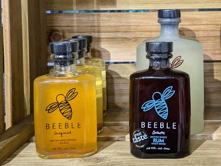 Three glass bottles of Beeble beverages are displayed on a wooden shelf. The leftmost bottle contains Beeble Original, a British honey whisky liqueur, with a golden-yellow liquid and a label featuring a bee illustration. The center bottle is a clear liqueur, while the rightmost bottle is labeled Beeble Swarm, a British honey rum spirit drink, showcasing a dark liquid and a prominent 