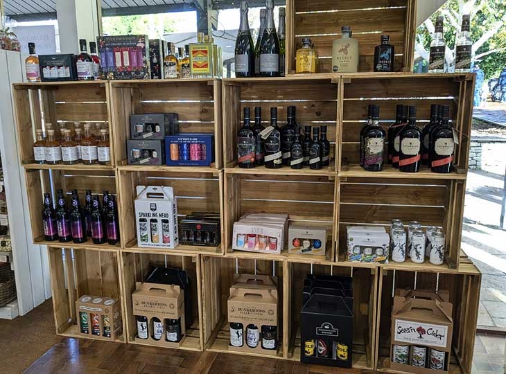 A wooden display shelf filled with various bottles and packaged beverages. The top shelves contain wine and spirits, including sparkling wine and liqueurs, while the middle shelves feature a selection of craft beers and meads. The bottom shelves display boxed sets and cans, showcasing a variety of drinks in an organized, rustic setting.